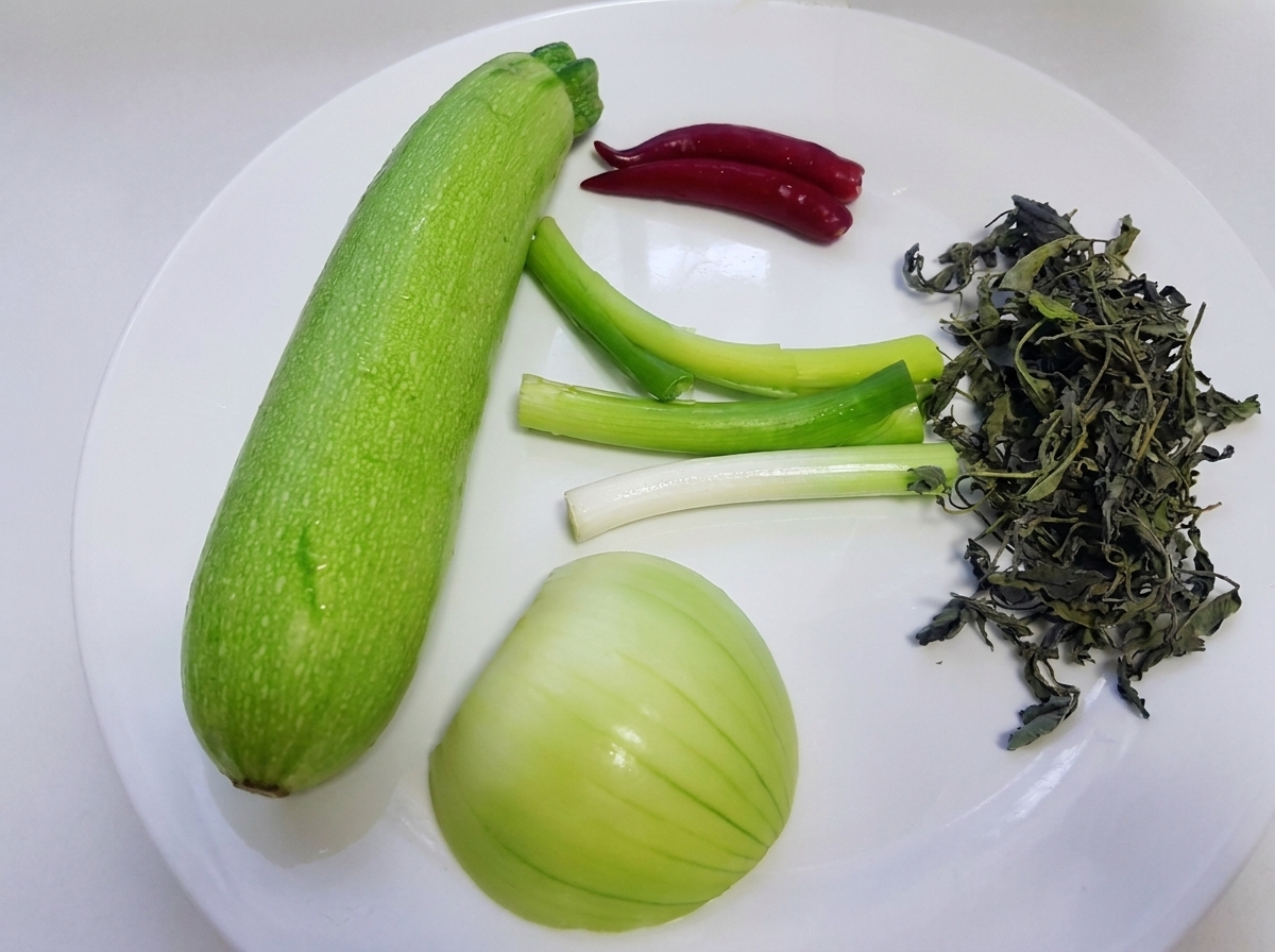 Surowe składniki, w tym cała zielona cukinia, szczypior, biała cebula, dzikie liście pachnotki i czerwone papryczki chili ułożone na białym talerzu.cite: 1