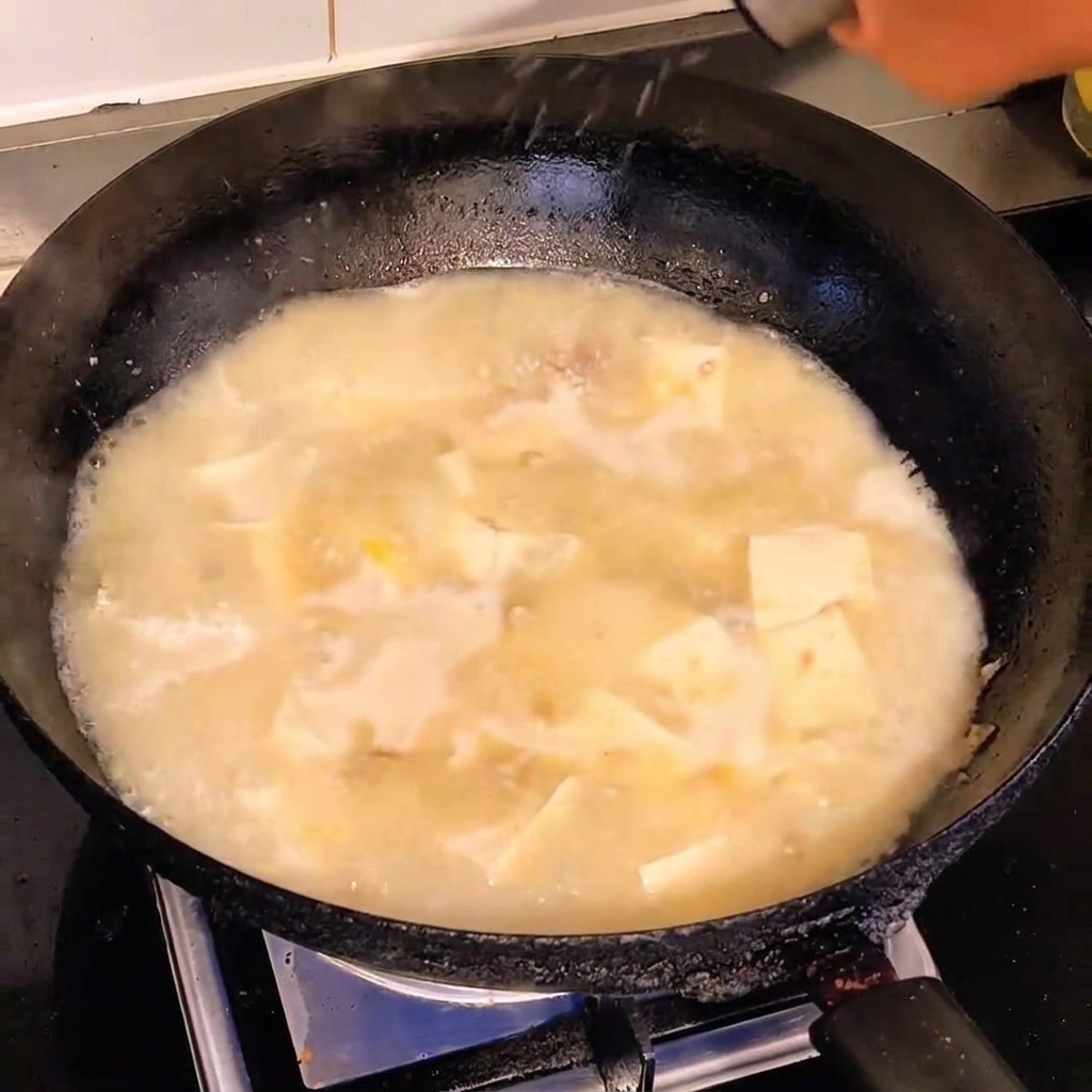 Zbliżenie na biały pieprz posypywany na bulgoczącą zupę z tofu i wieprzowiną w ciemnym woku.