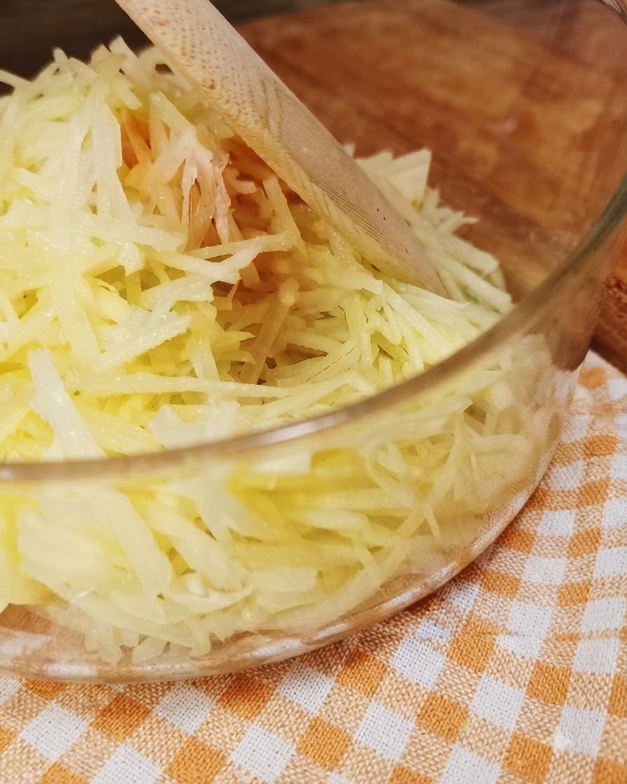 Świeżo starte ziemniaki wewnątrz przezroczystej szklanej miski z drewnianym przyborem.