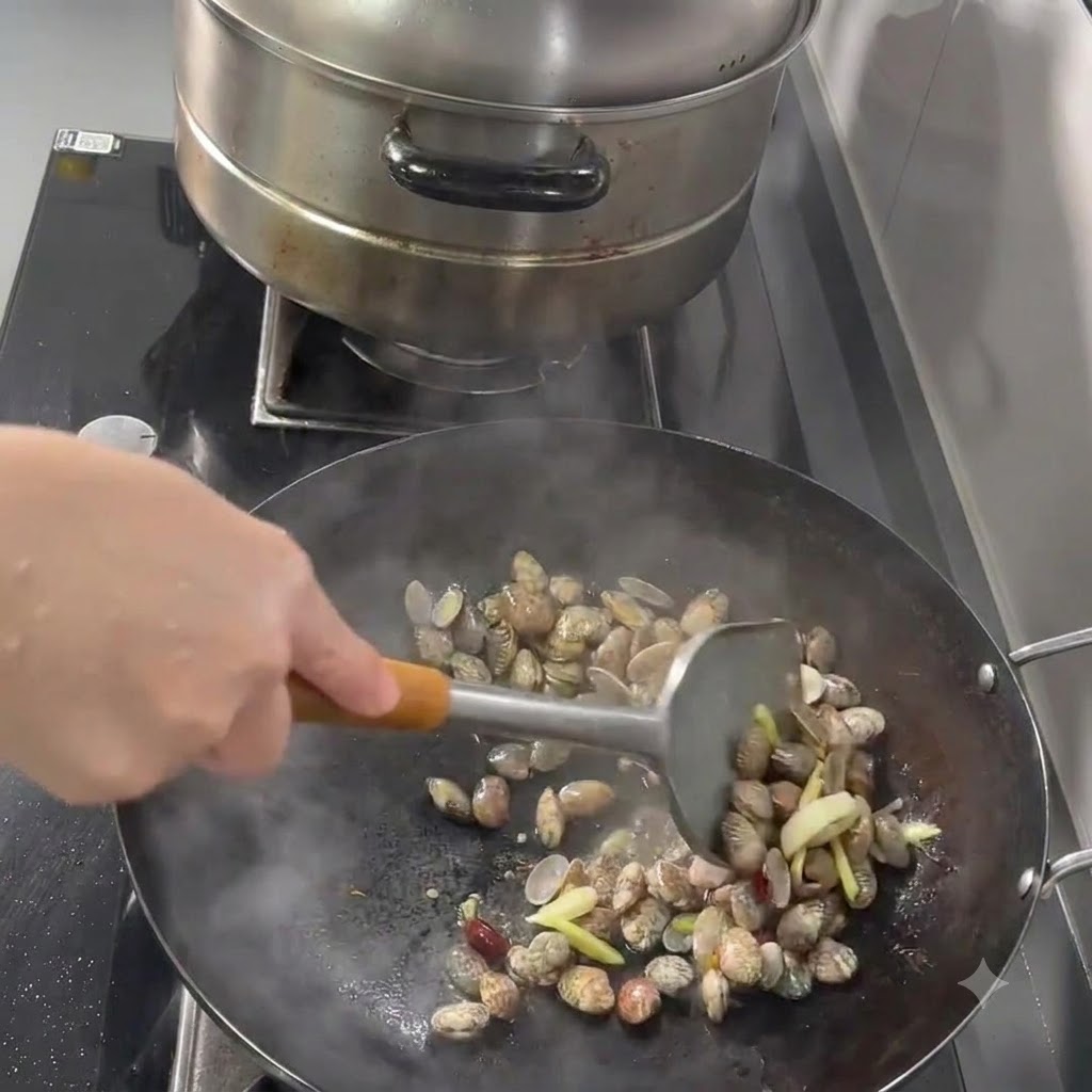 Małże, czosnek, imbir i chili smażone w woku za pomocą metalowej łopatki.