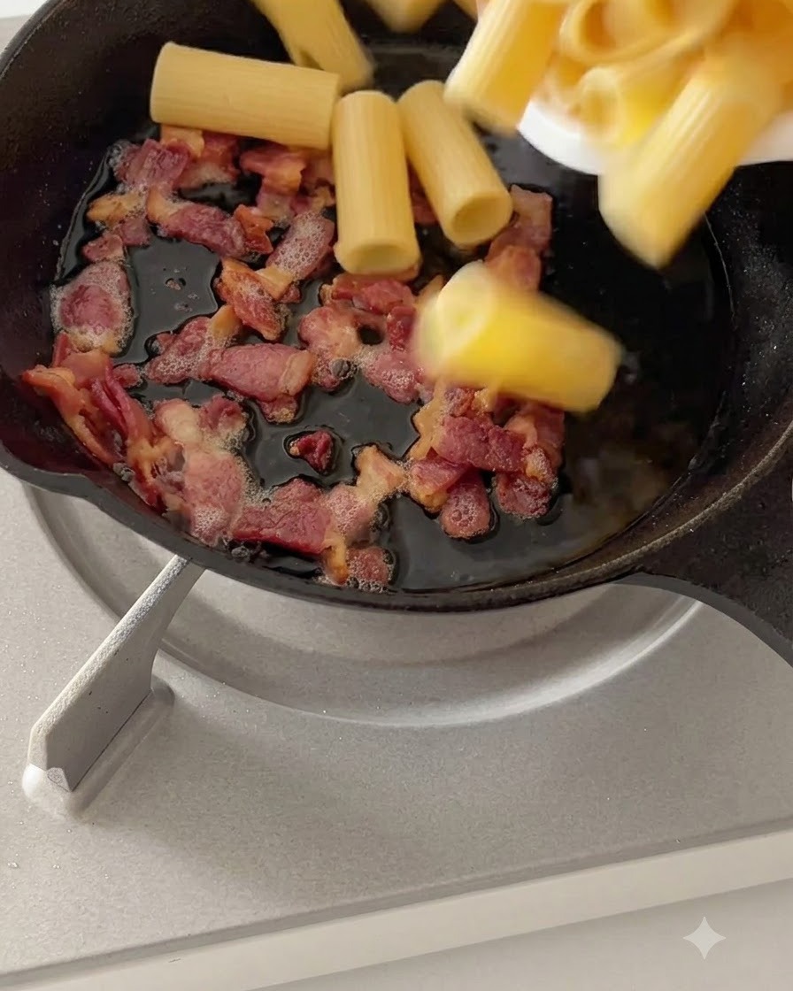 Ugotowany makaron rigatoni mieszany z usmażonym boczkiem na patelni na małym ogniu.
