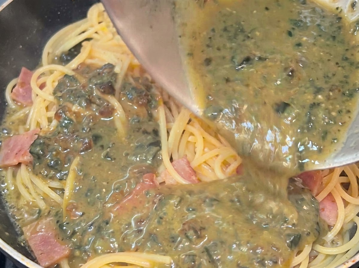 Kremowy, nakrapiany sos z jaj wiekowych wylewany z miski na gorące spaghetti i boczek na patelni.
