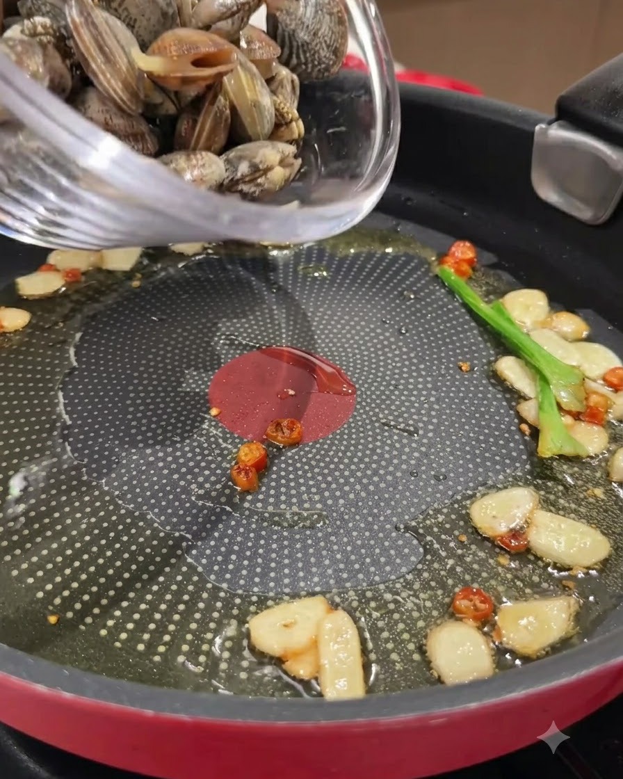 Świeże małże wsypywane ze szklanej miski na patelnię z podsmażonym czosnkiem, chilli i oliwą.