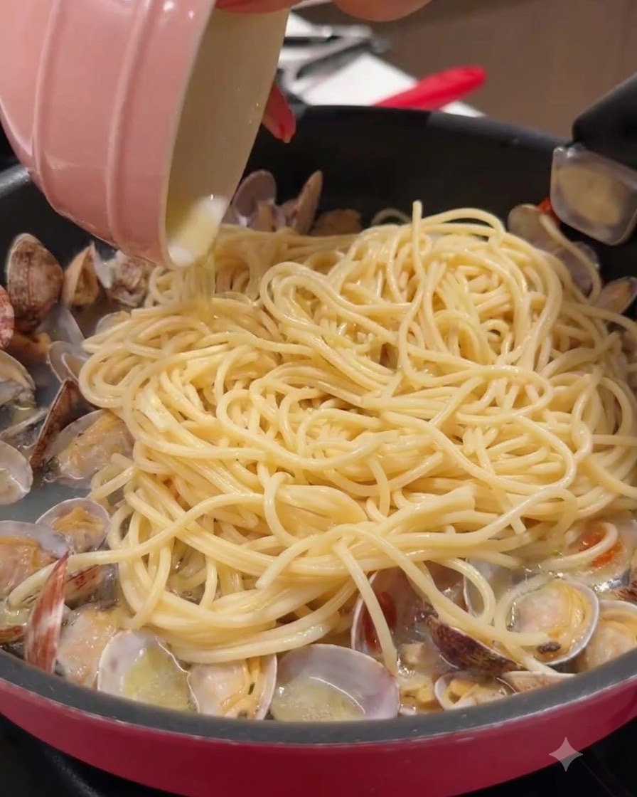 Mała różowa miseczka wlewająca płyn na patelnię wypełnioną spaghetti i małżami.