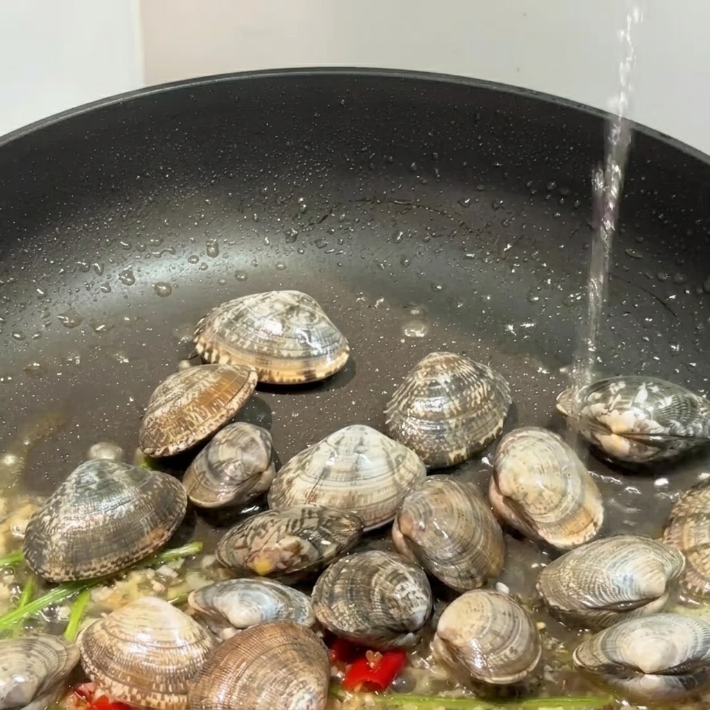 Czarna patelnia z małżami, czosnkiem i chili w momencie dolewania płynu, przygotowywana do przykrycia.