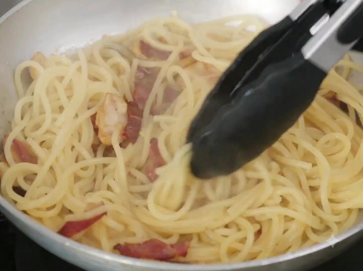 Masło i woda z makaronu mieszane ze spaghetti na patelni za pomocą szczypiec.