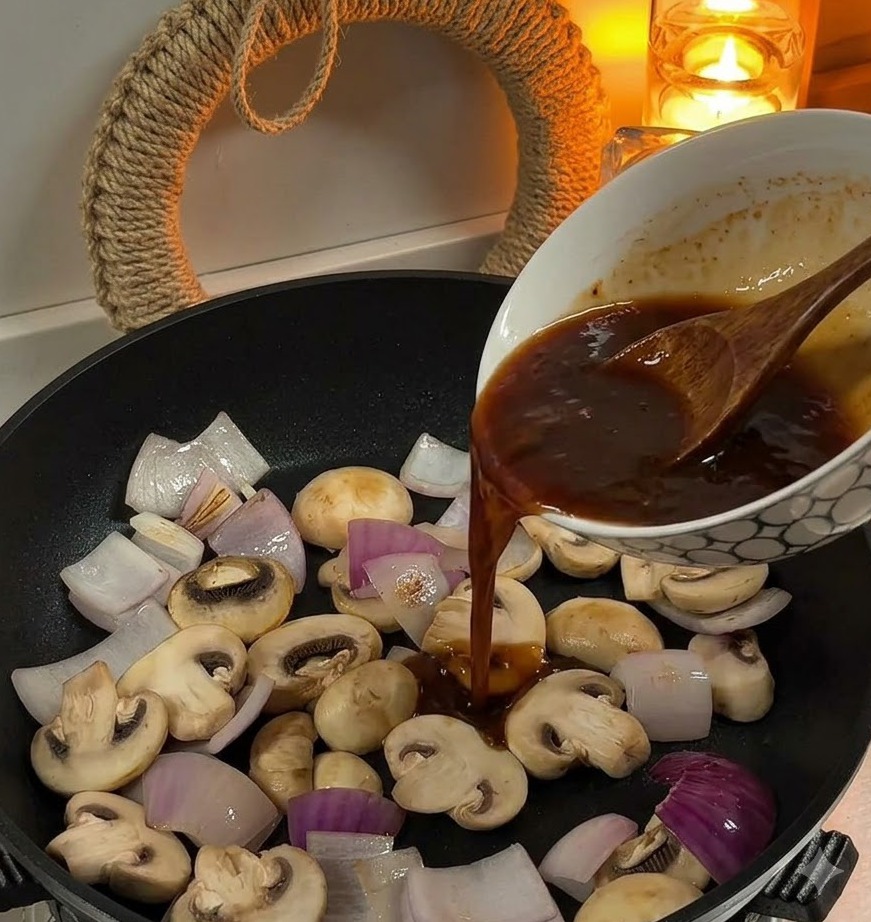 Wlewanie brązowego sosu z miski na patelnię zawierającą pokrojoną cebulę i białe pieczarki.