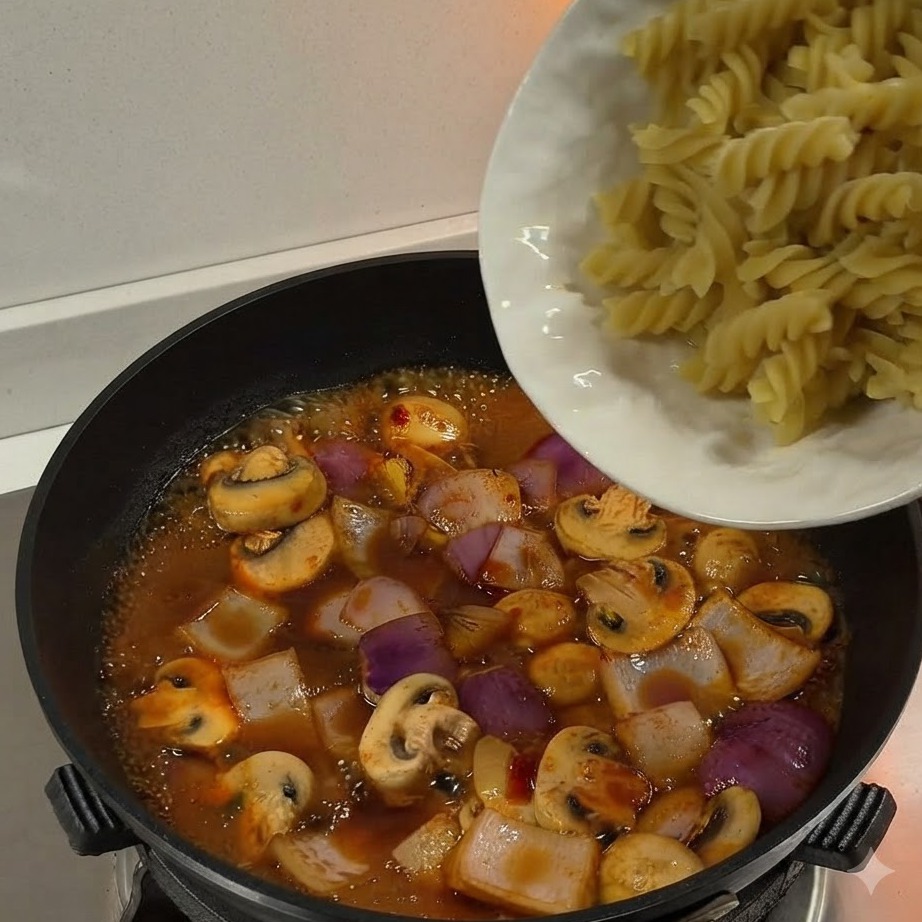 Ugotowany makaron fusilli przekładany z białego talerza na patelnię pełną duszącego się sosu i warzyw.