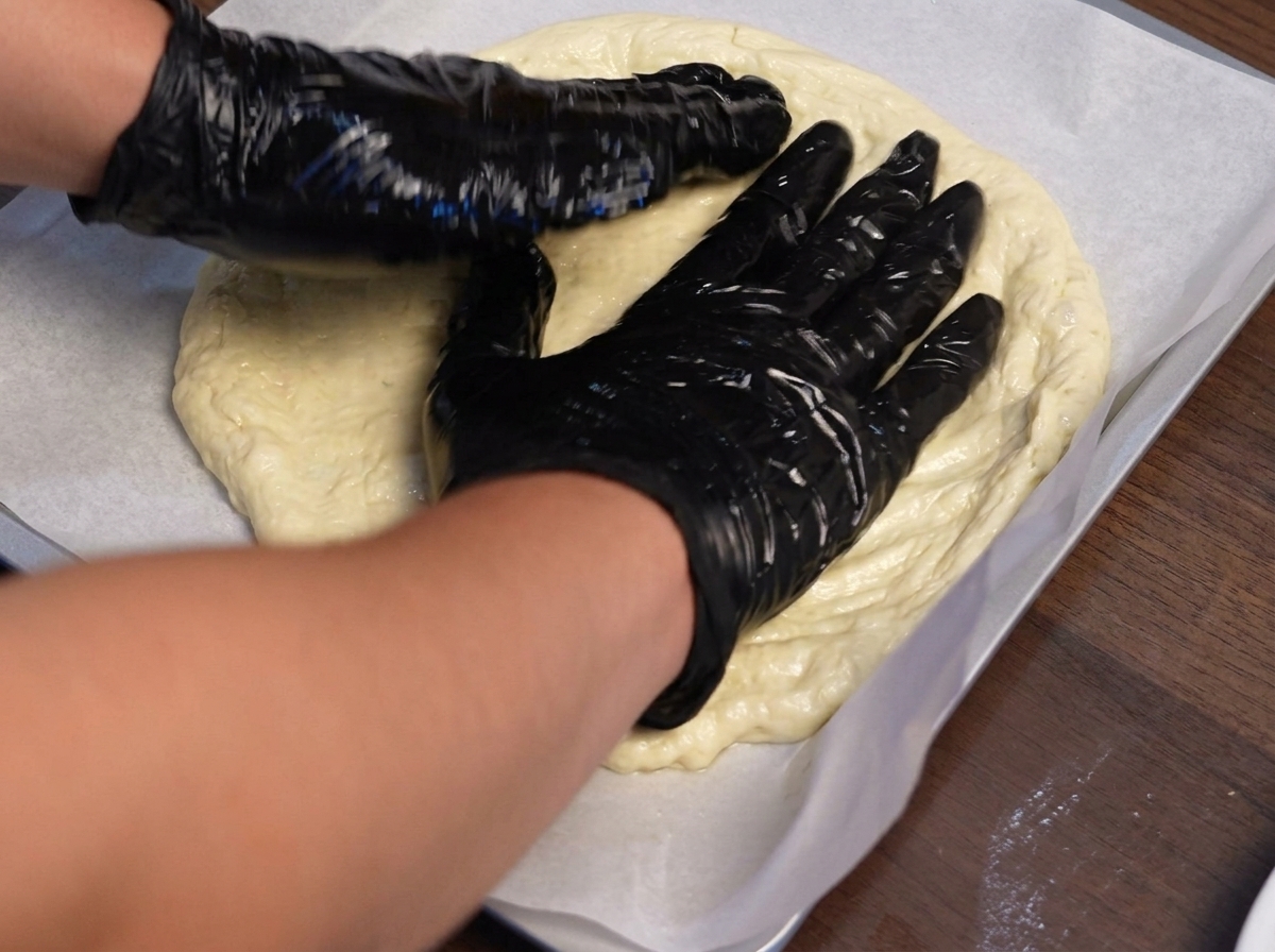 Ręce w rękawiczkach kształtujące ciasto na pizzę na blaszce wyłożonej papierem do pieczenia.