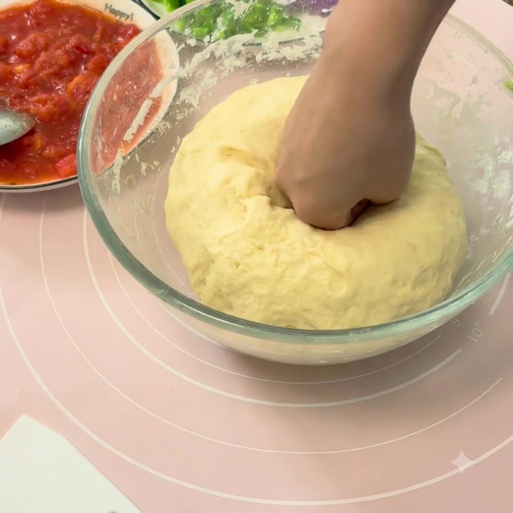 Dłoń wciskająca do wnętrza dużą kulę wyrośniętego ciasta na pizzę w szklanej misce.
