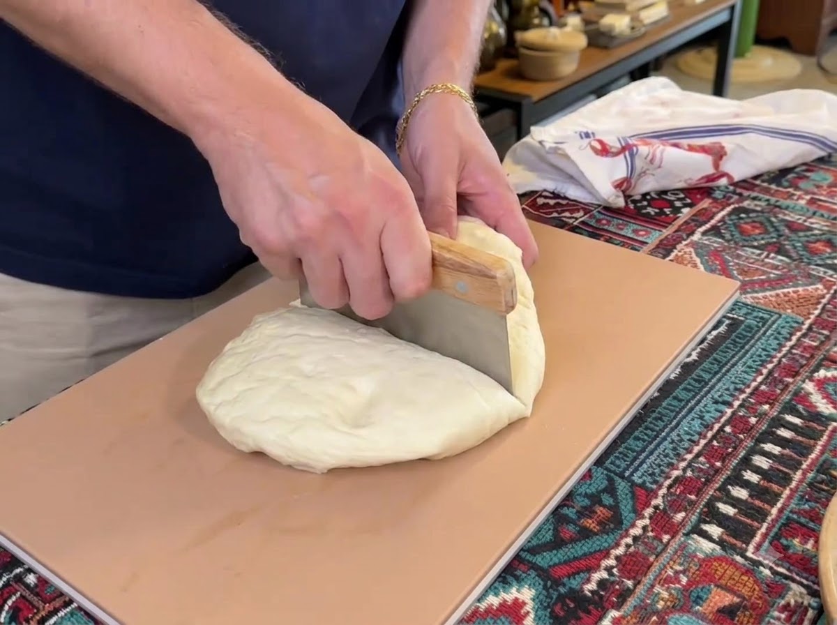 Skrobka do ciasta krojąca dużą grudę białego ciasta na pizzę na cztery oddzielne części na desce do krojenia.