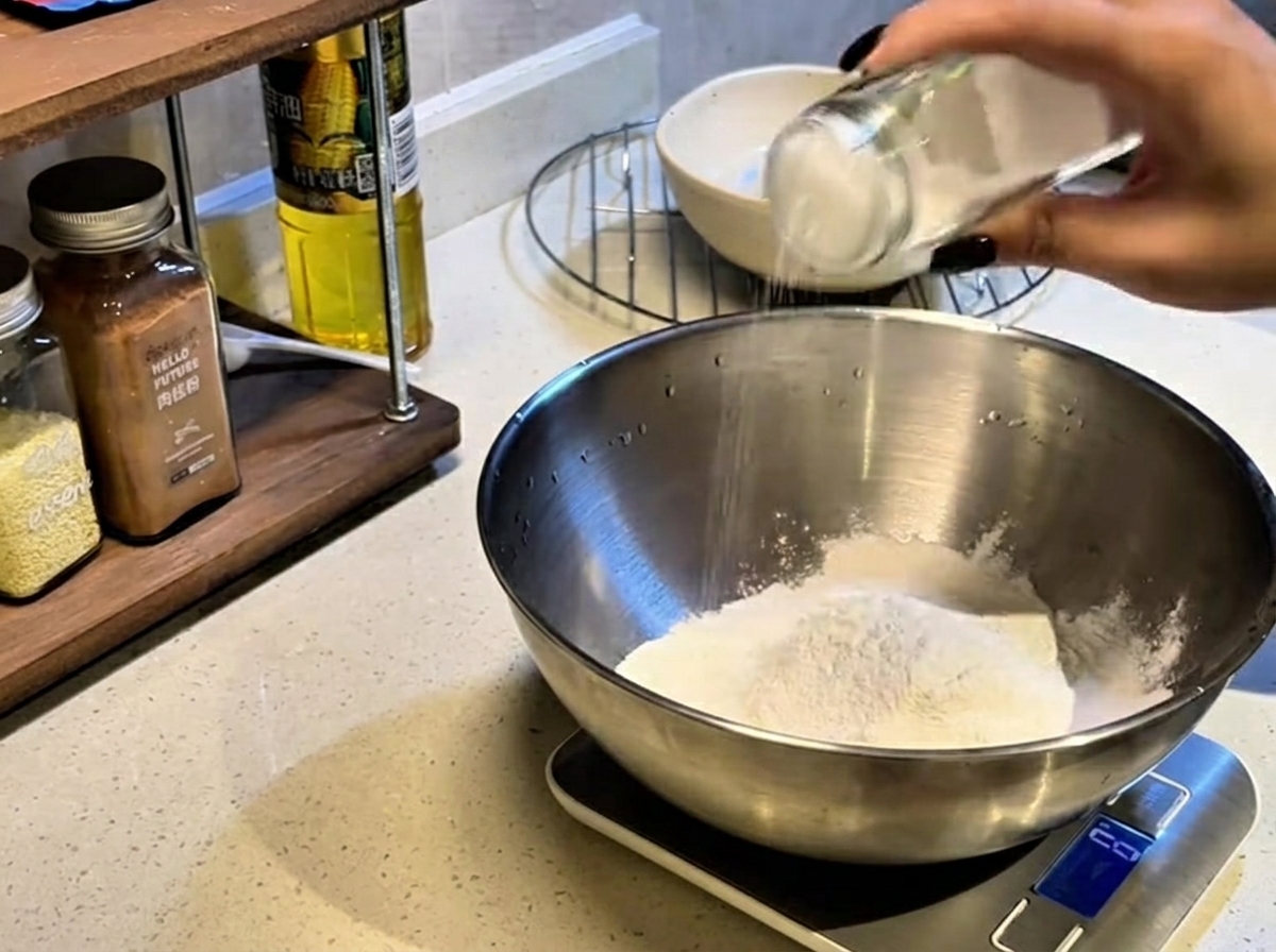 Ręka potrząsająca szklanym słoikiem w celu posypania solą kopczyka białej mąki w dużej metalowej misce.