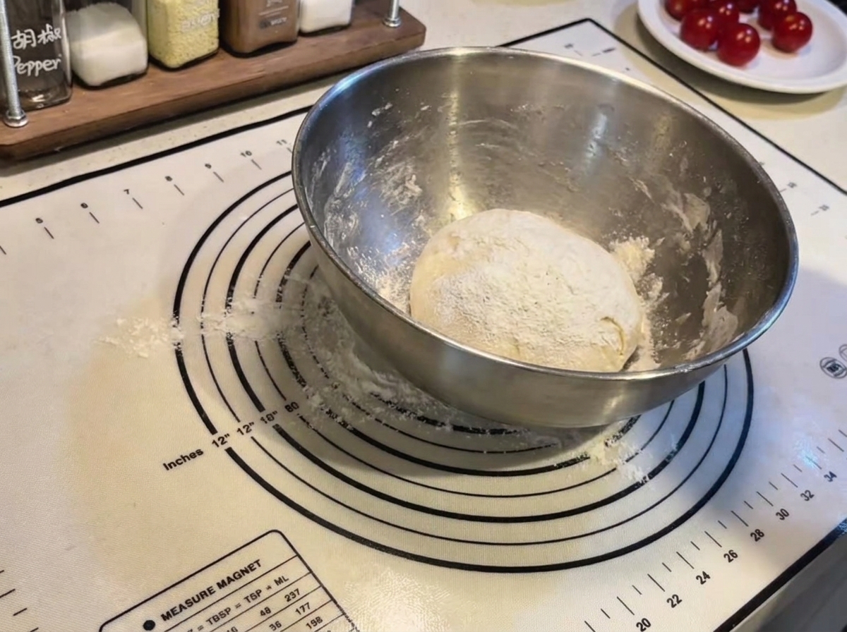 Okrągła kula ciasta do pizzy oprószona mąką wewnątrz stalowej miski na silikonowej macie do pieczenia.