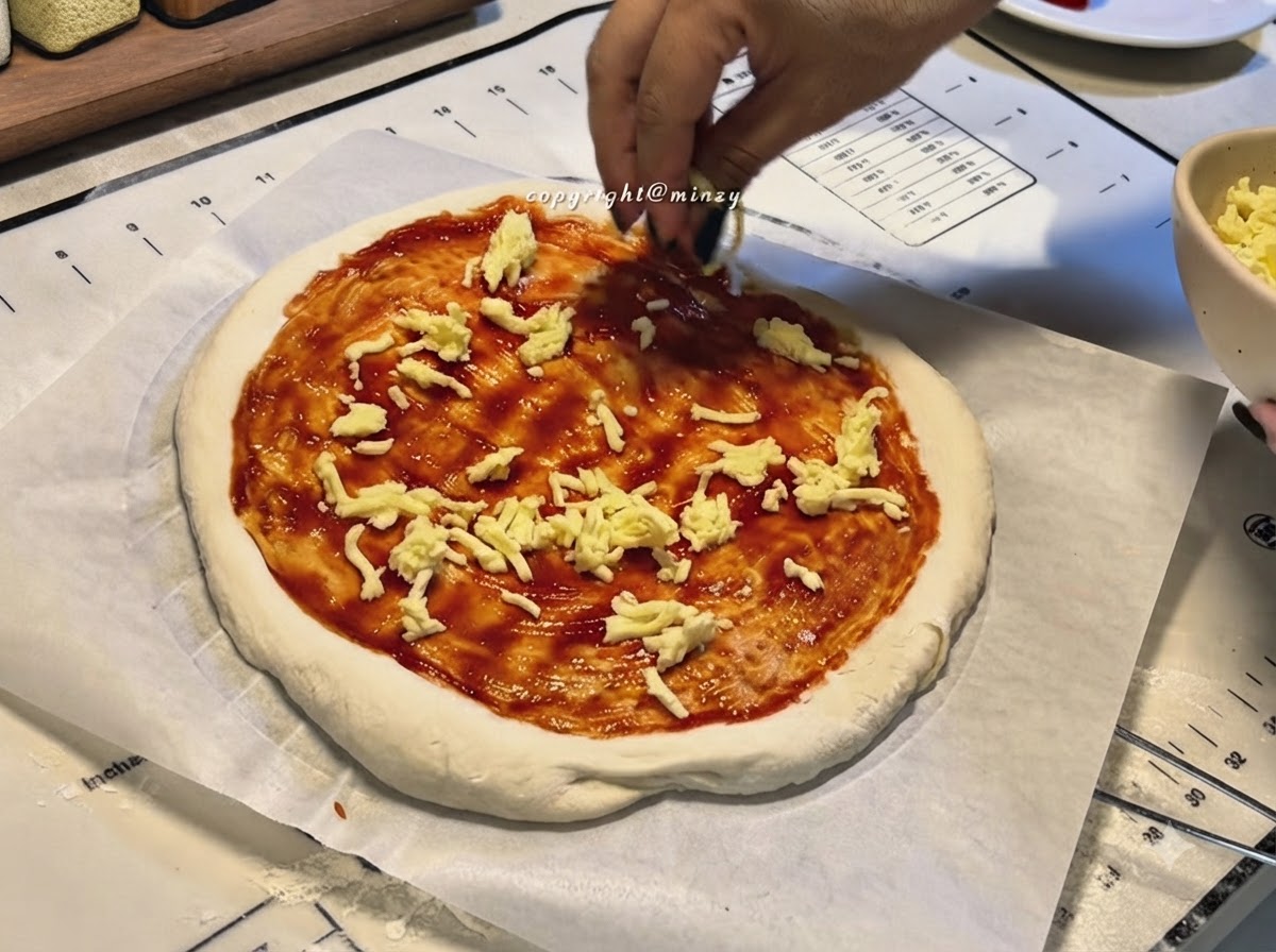 Dłonie posypujące startym serem spód pizzy pokryty czerwonym sosem.
