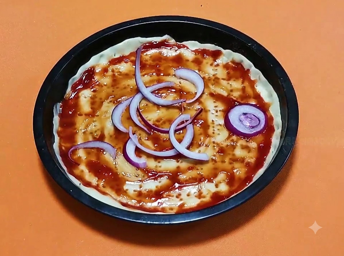 Krążki czerwonej cebuli ułożone na bazie ciasta do pizzy z sosem pomidorowym wewnątrz czarnej, okrągłej formy do pieczenia.