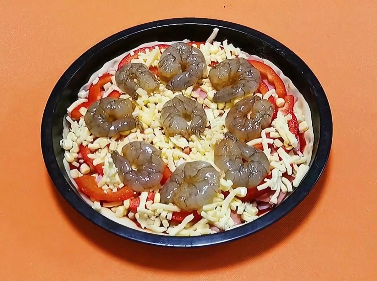 Surowe zamarynowane krewetki ułożone w okrąg na wierzchu pizzy z serem, papryką i szynką.