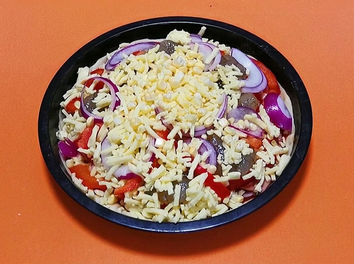 W pełni uformowana surowa pizza z owocami morza posypana ostatnią warstwą tartego sera i krążkami czerwonej cebuli, gotowa do pieczenia.