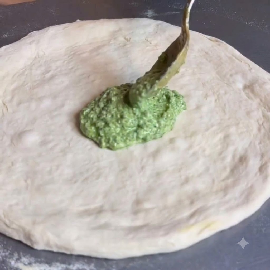 Łyżka rozprowadzająca zielone pesto na środku płaskiego, okrągłego ciasta na pizzę.