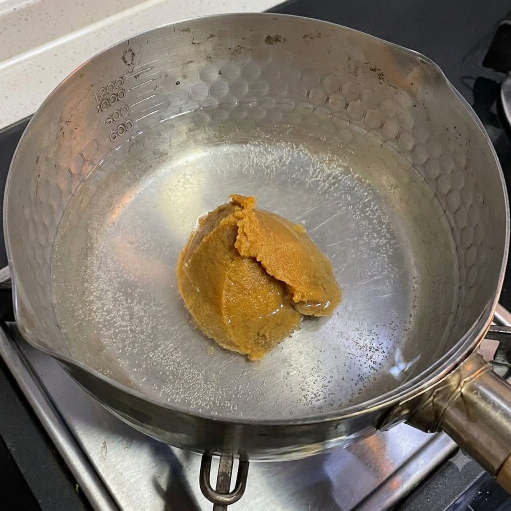 Łyżka białej pasty miso rozpuszczająca się w garnku z gorącą wodą na kuchence.