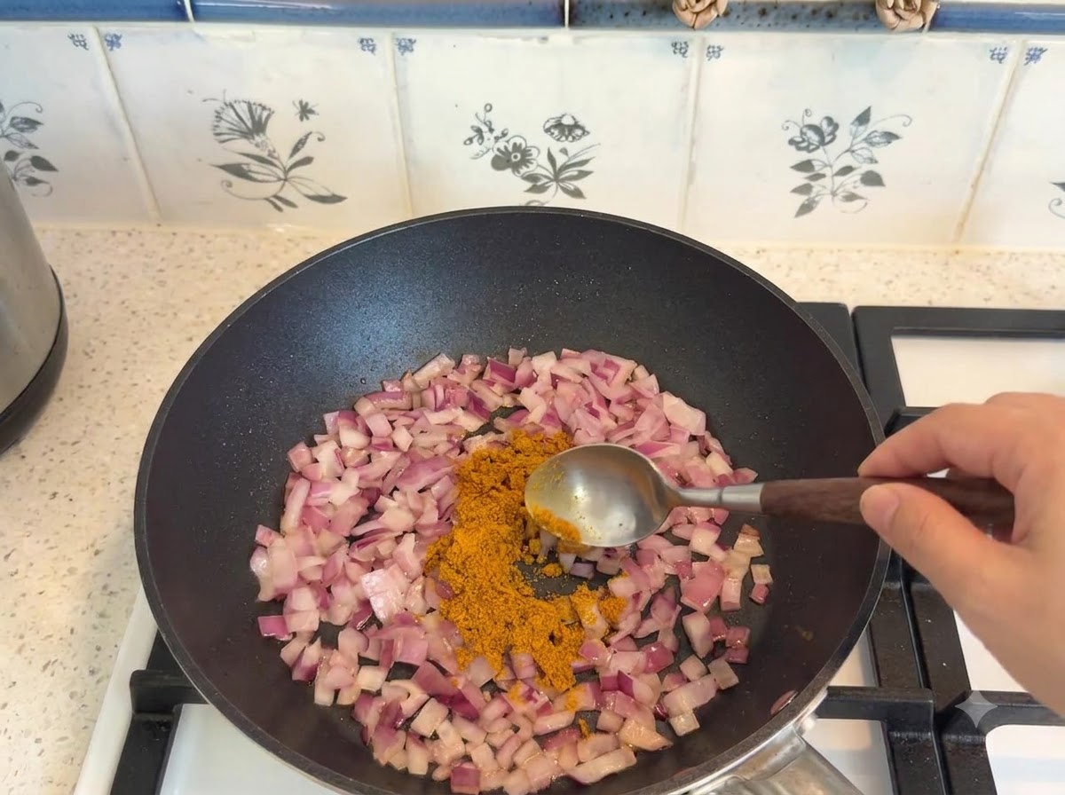 Przyprawa curry odmierzana łyżką i dodawana do podsmażonej cebuli na patelni.