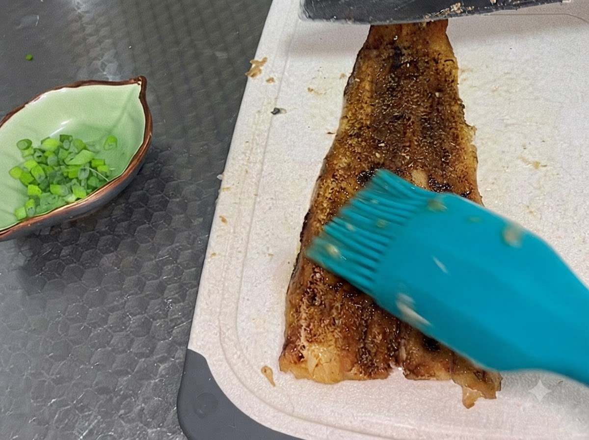 W pełni upieczony filet z węgorza krojony na białej desce obok małej, zielonej miseczki wypełnionej posiekaną dymką.