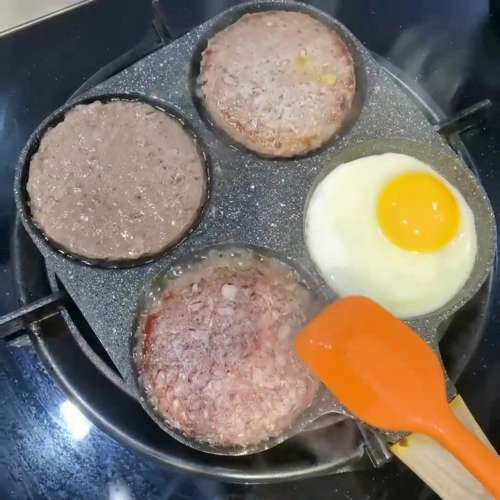 Kotlety burgerowe odwracane pomarańczową łopatką obok smażącego się jajka w sekcji patelni.
