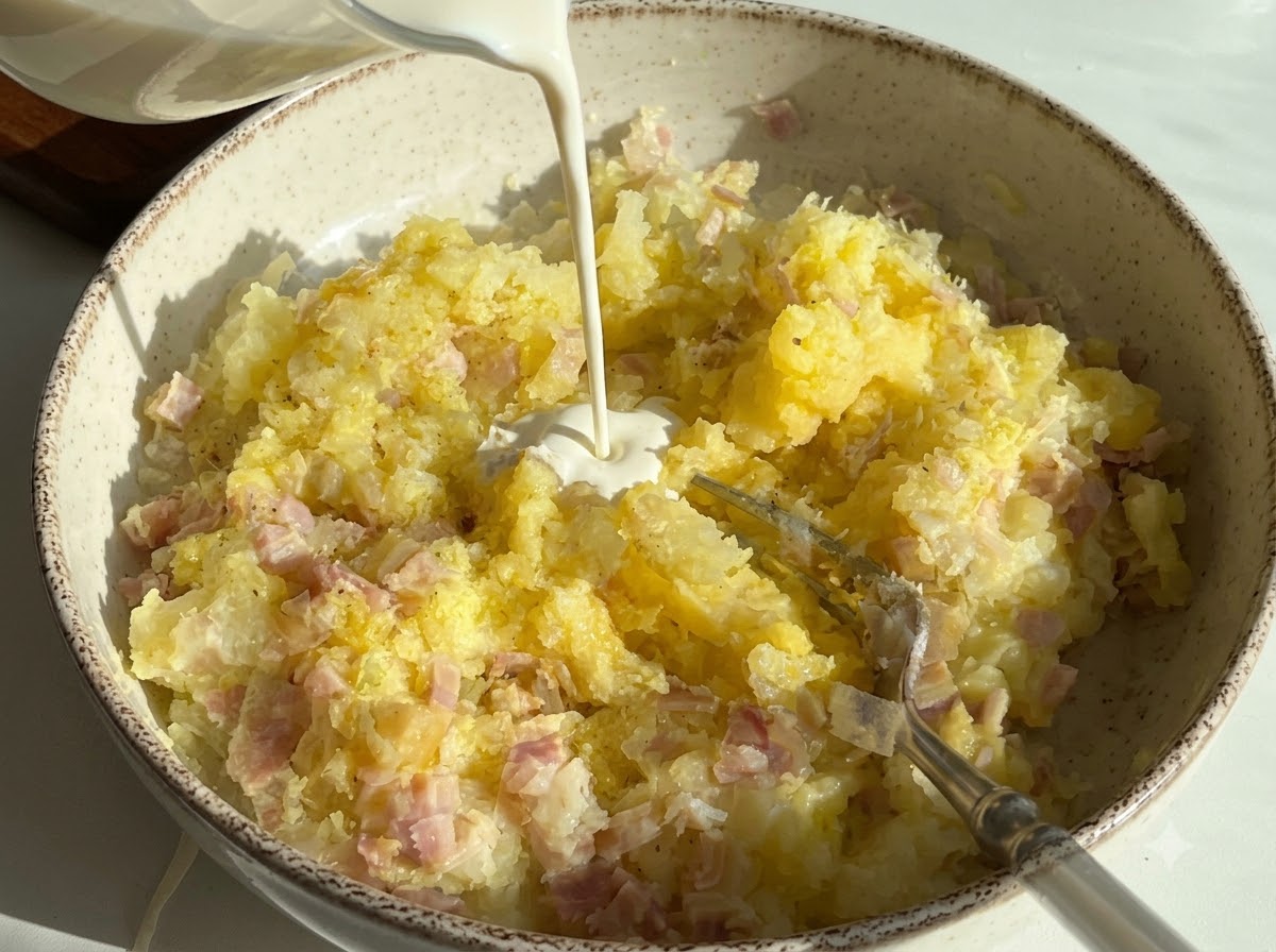 Śmietanka kremówka wlewana do miski zawierającej puree ziemniaczane i pokrojoną szynkę.
