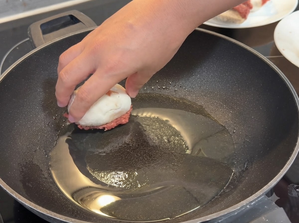Dłoń kładąca faszerowaną pieczarkę mięsem do dołu na rozgrzanej patelni z olejem.