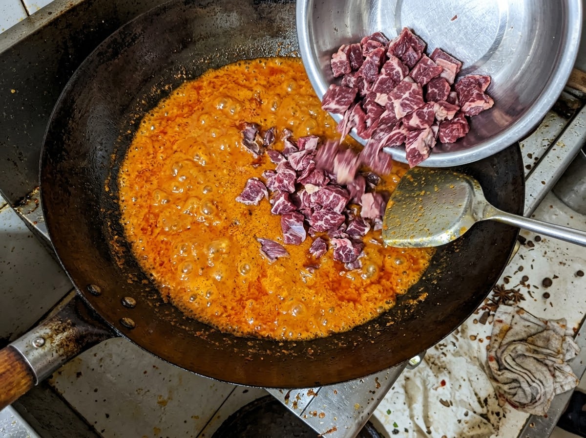 Kawałki wołowiny przekładane z metalowej miski do bulgoczącej czerwonej bazy chili w woku.
