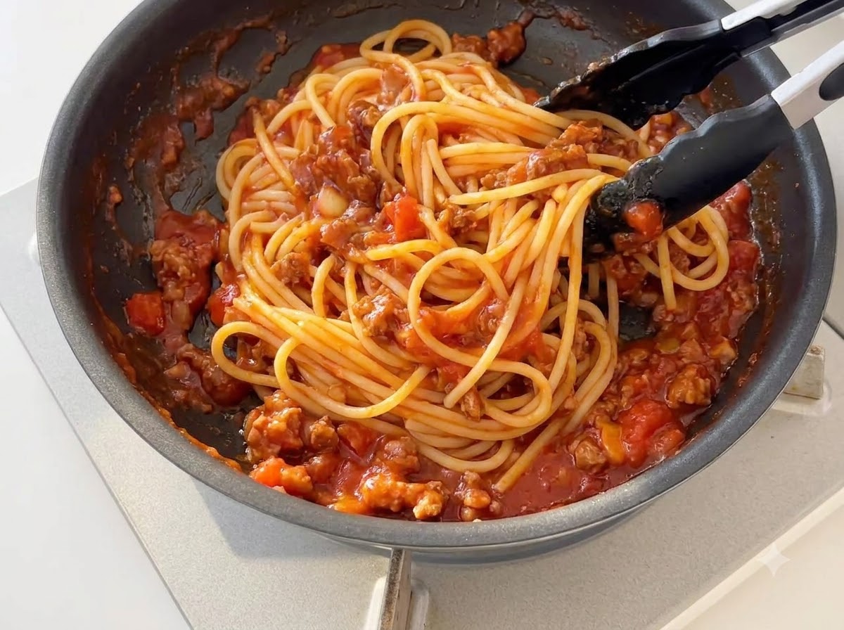 Ugotowany makaron spaghetti mieszany z zagęszczonym pomidorowym sosem mięsnym na patelni za pomocą czarnych silikonowych szczypców.