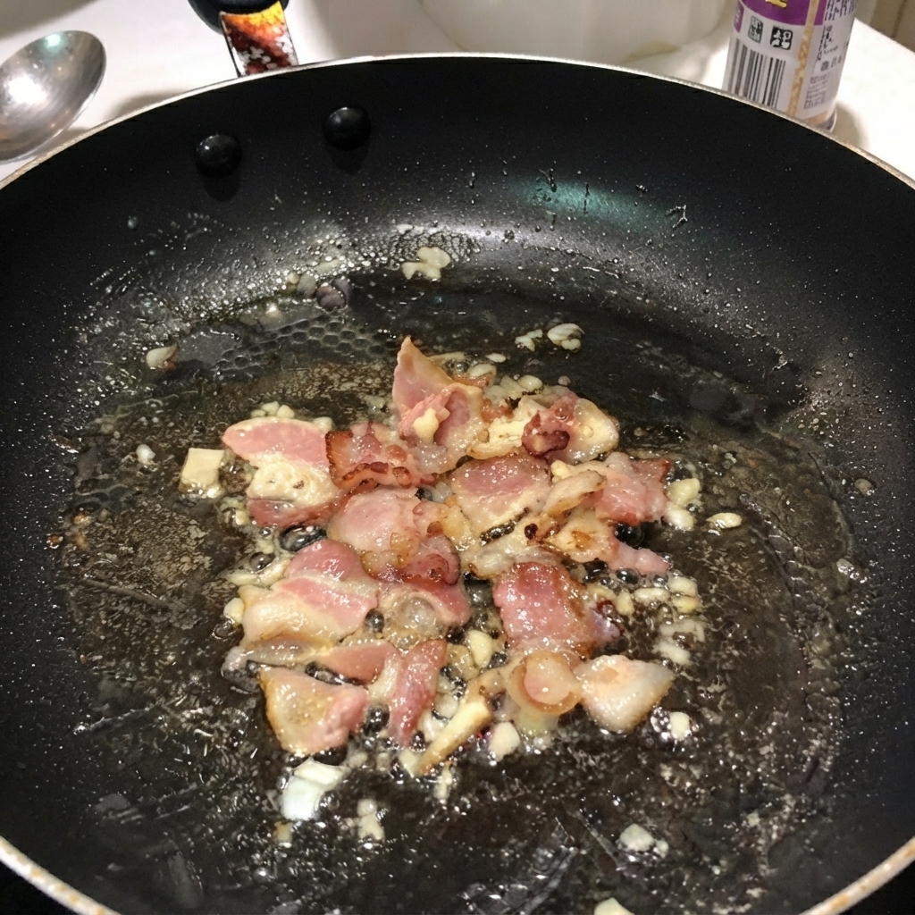 Ugotowany makaron penne wymieszany z boczkiem i cebulą na czarnej patelni.