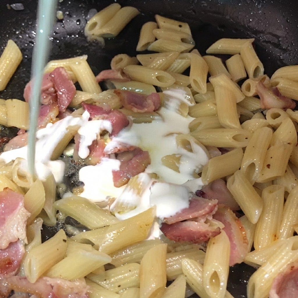 Mleko i śmietanka wlewane od góry do patelni wypełnionej makaronem penne i kawałkami boczku.