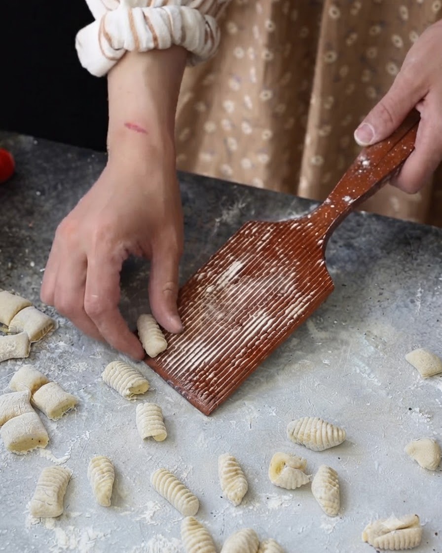 Ręka rolująca mały kawałek ciasta po drewnianej, rowkowanej desce do gnocchi.
