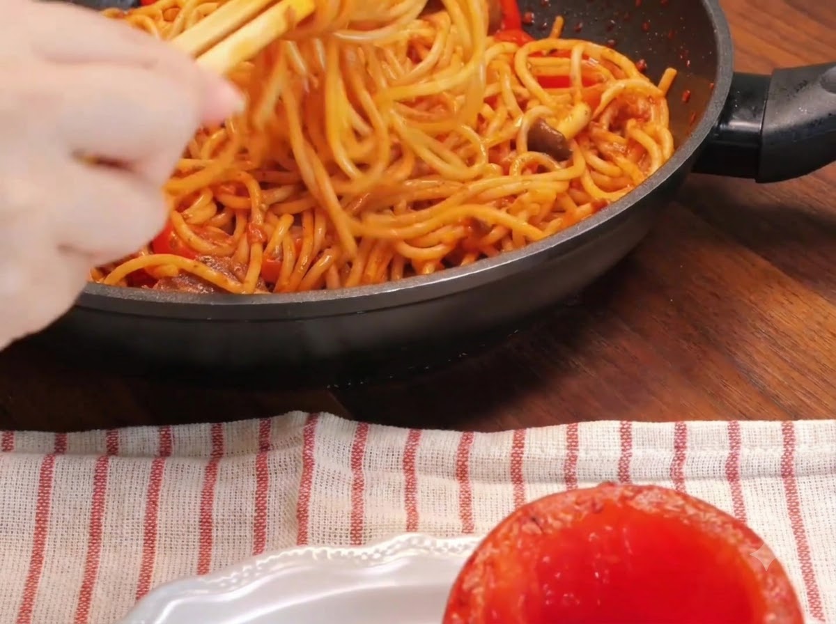 Zbliżenie dłoni używającej pałeczek do kręcenia klusek spaghetti na patelni obok wydrążonego, pieczonego pomidora na talerzu.