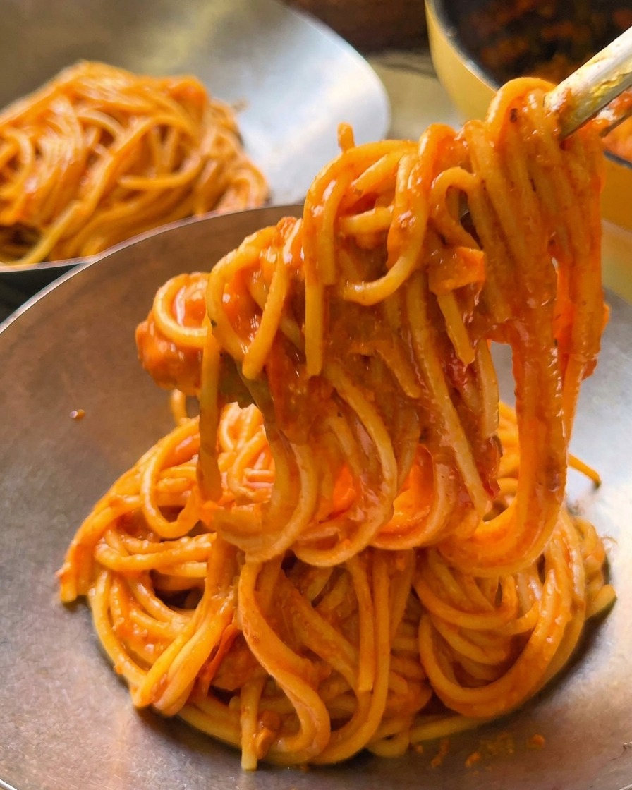 Szczypce unoszące porcję kremowego spaghetti pomidorowego do polerowanej metalowej miski do serwowania.