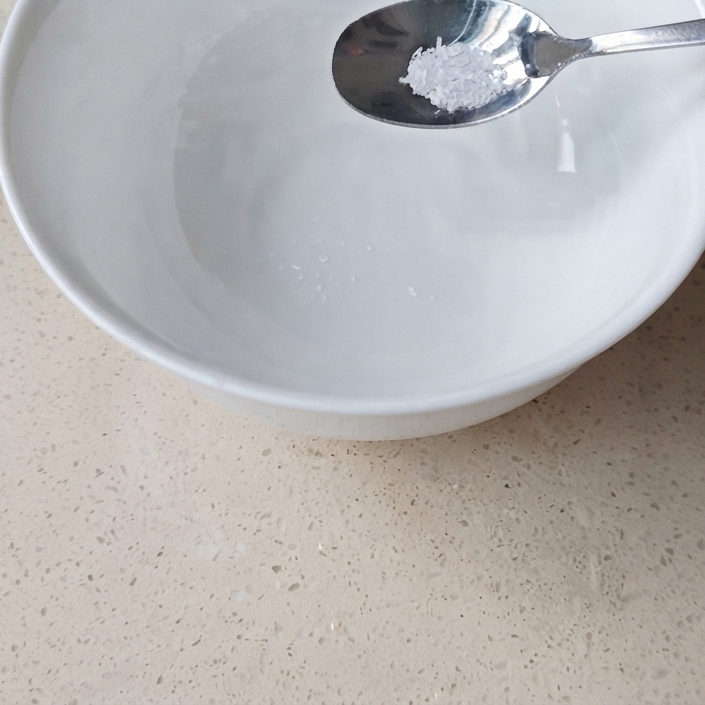 Metalowa łyżka z niewielką ilością glutaminianu sodu trzymana nad białą, ceramiczną miską.