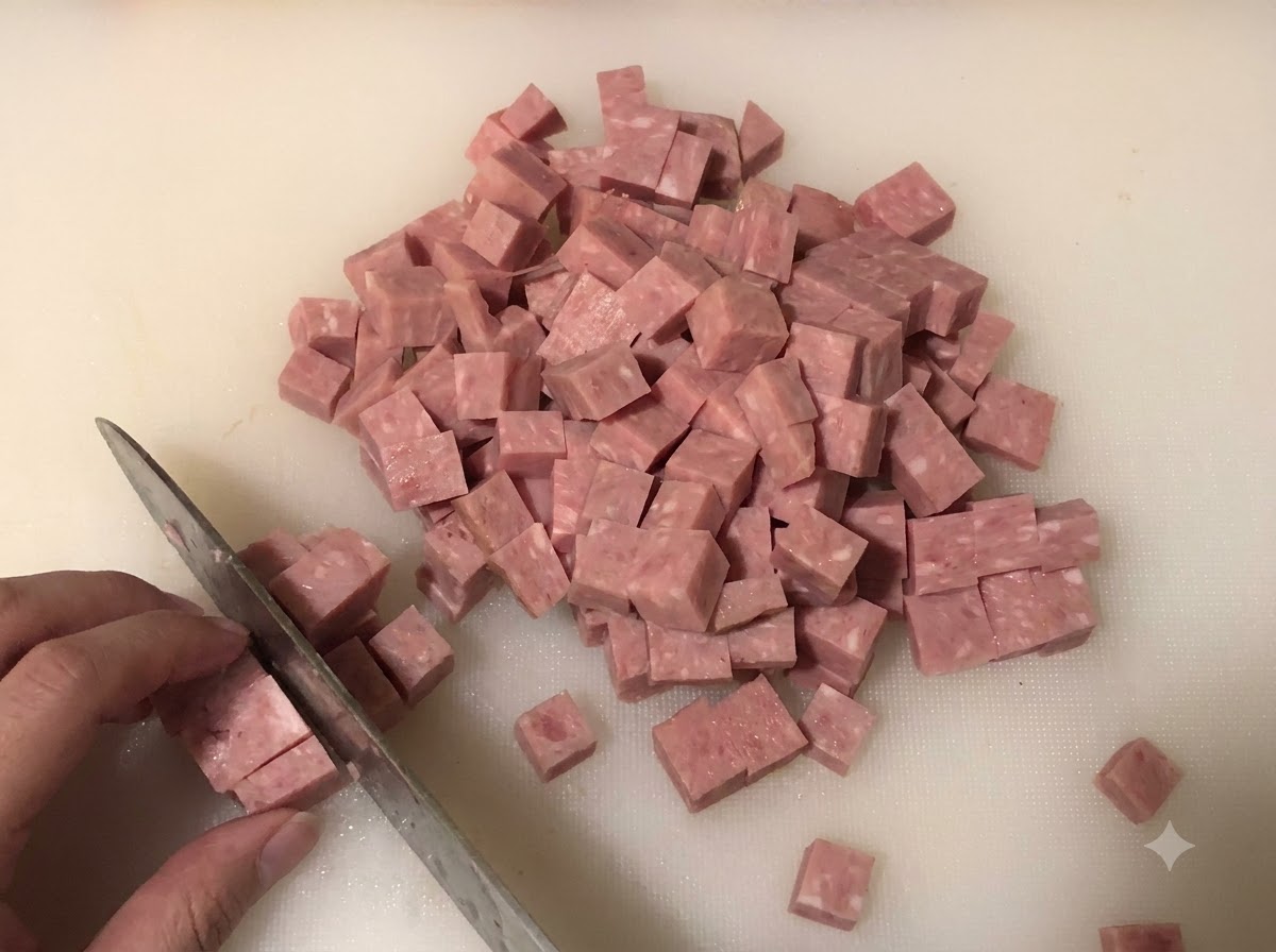 Spam pokrojony w małą kostkę na białej desce do krojenia, w tle posiekana cebula i zielona cebulka.
