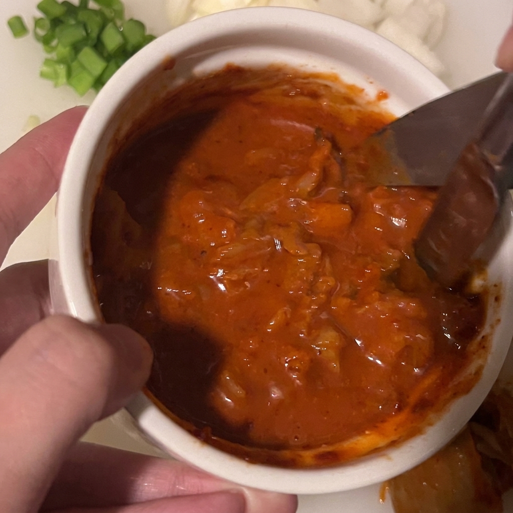 Nożyczki kuchenne tnące kawałki kimchi zanurzone w gęstym czerwonym sosie w małej białej misce.