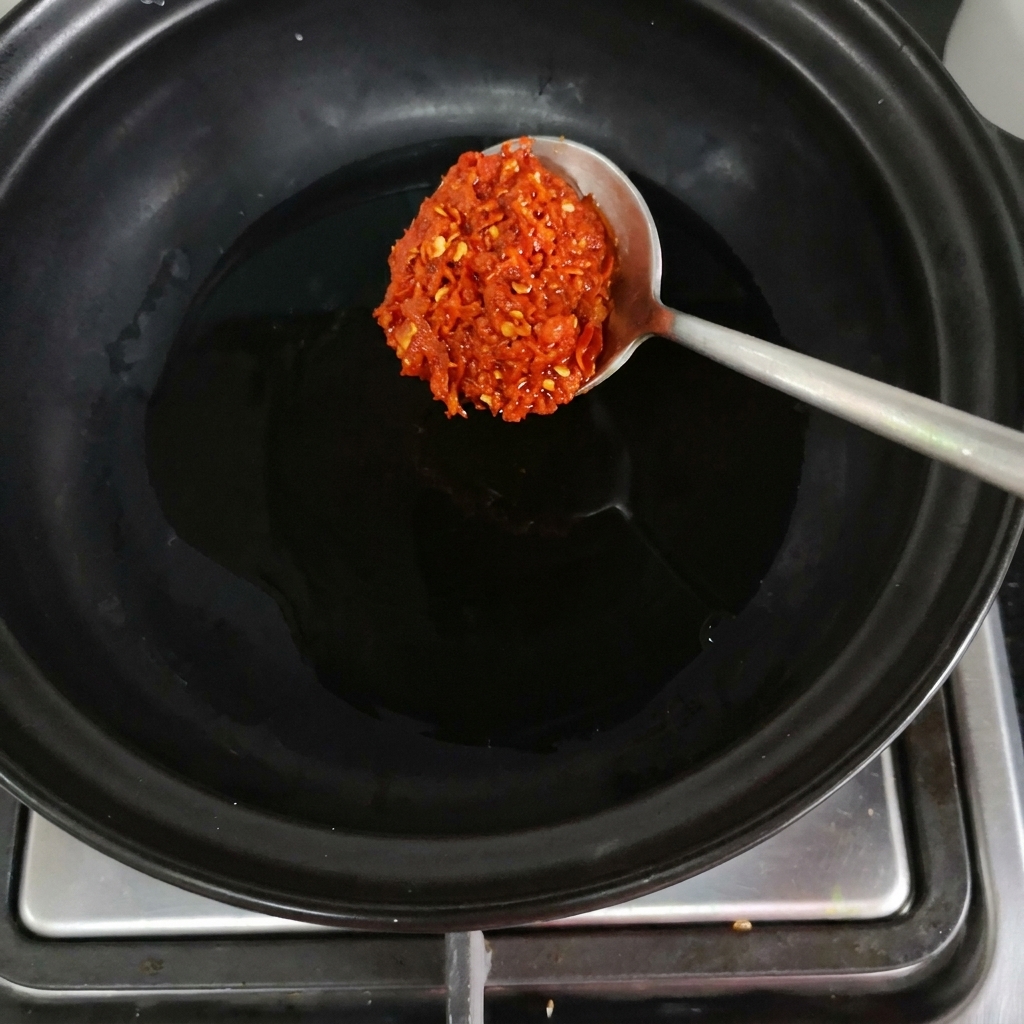 Łyżka czerwonej pasty chili wrzucana do czarnego garnka glinianego na palniku.