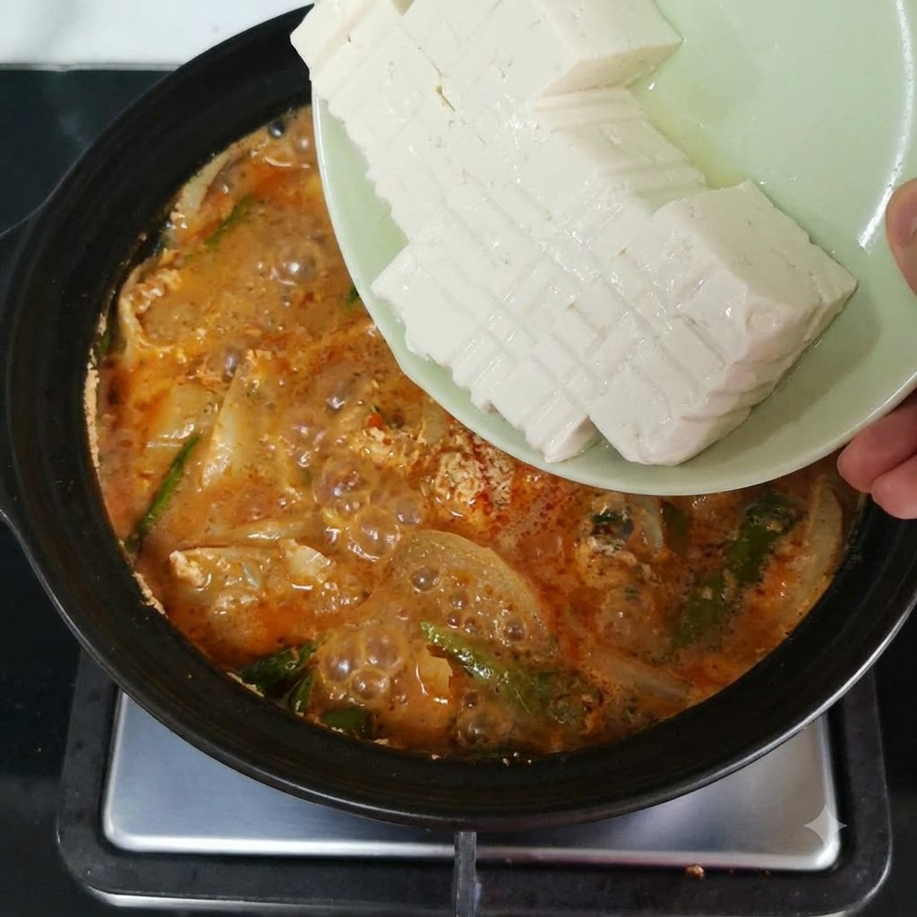 Pokrojone miękkie tofu zsuwane z talerza do gotującego się garnka wypełnionego pomarańczowo-czerwonym bulionem i warzywami.