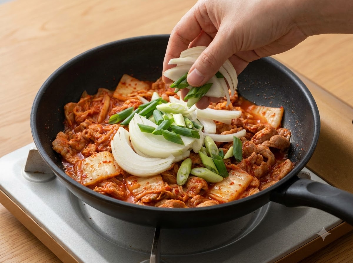 Pokrojona w plasterki cebula i zielona cebulka dodawane do garnka na warstwę kimchi.