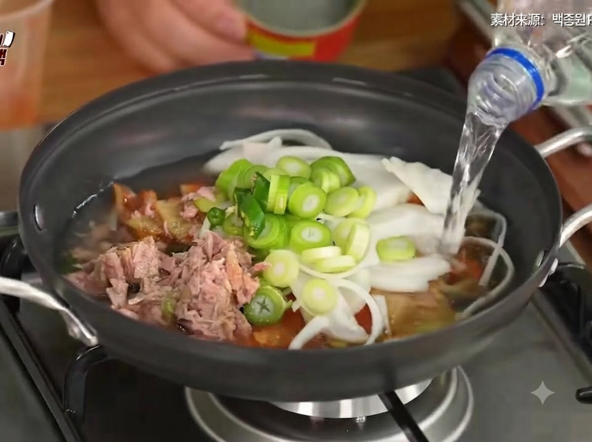 Woda wlewana do garnka zawierającego kimchi, warzywa i tuńczyka.