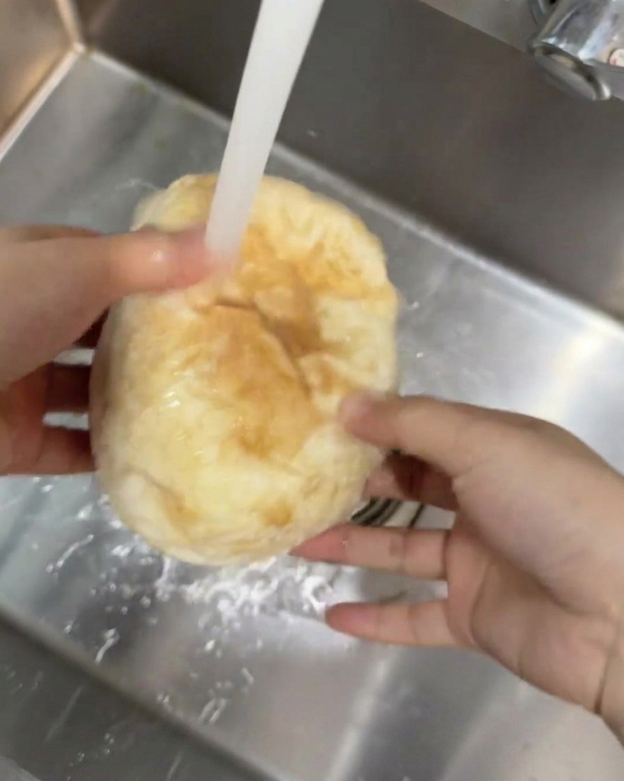 Bochenek chleba ciabatta trzymany pod strumieniem bieżącej wody w zlewie kuchennym.