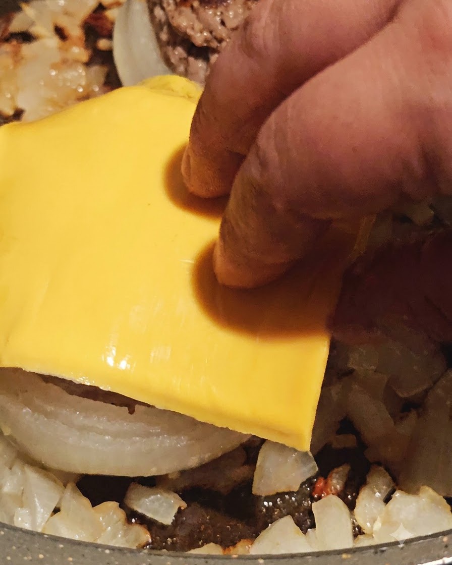 Plaster żółtego sera kładziony na wierzchu usmażonego krążka cebulowego z mięsem na patelni.