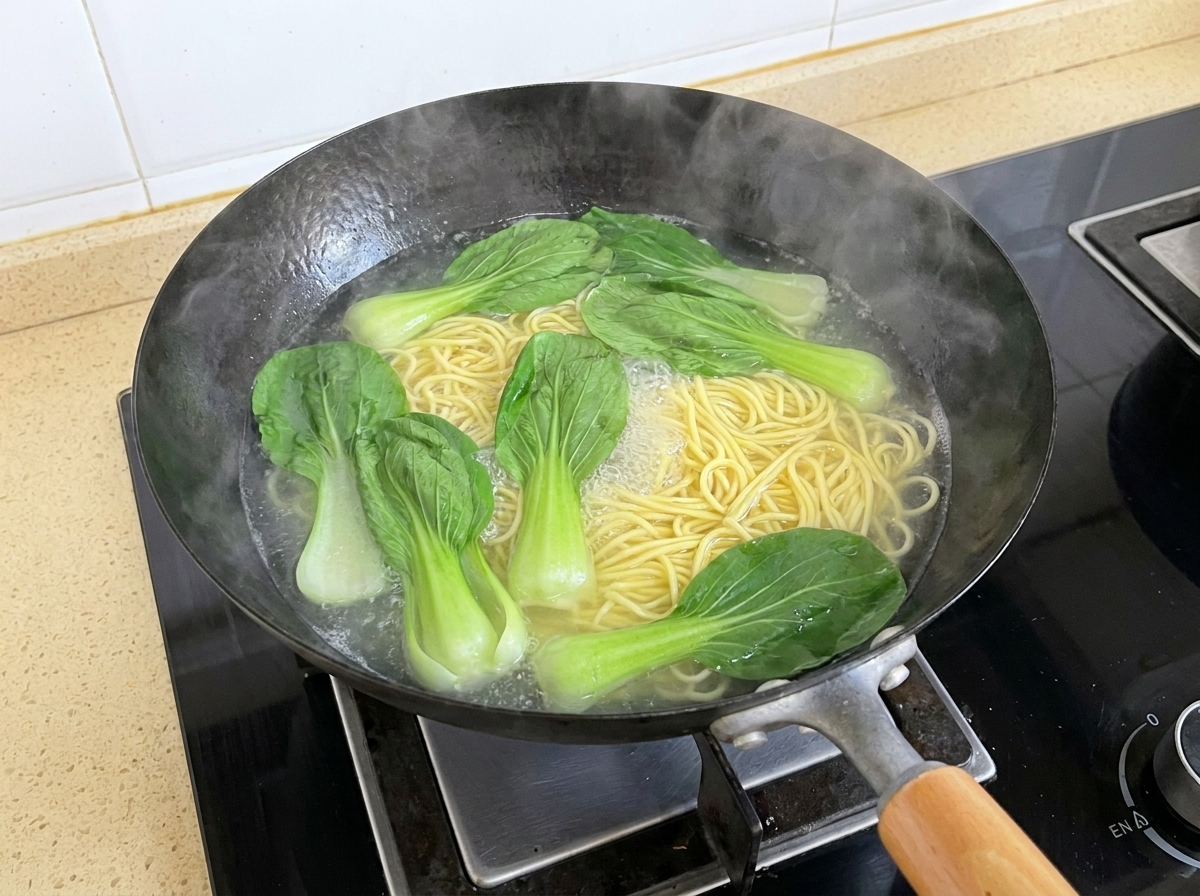 Świeże liście kapusty bok choy pływające we wrzącej wodzie obok gotującego się makaronu w woku.