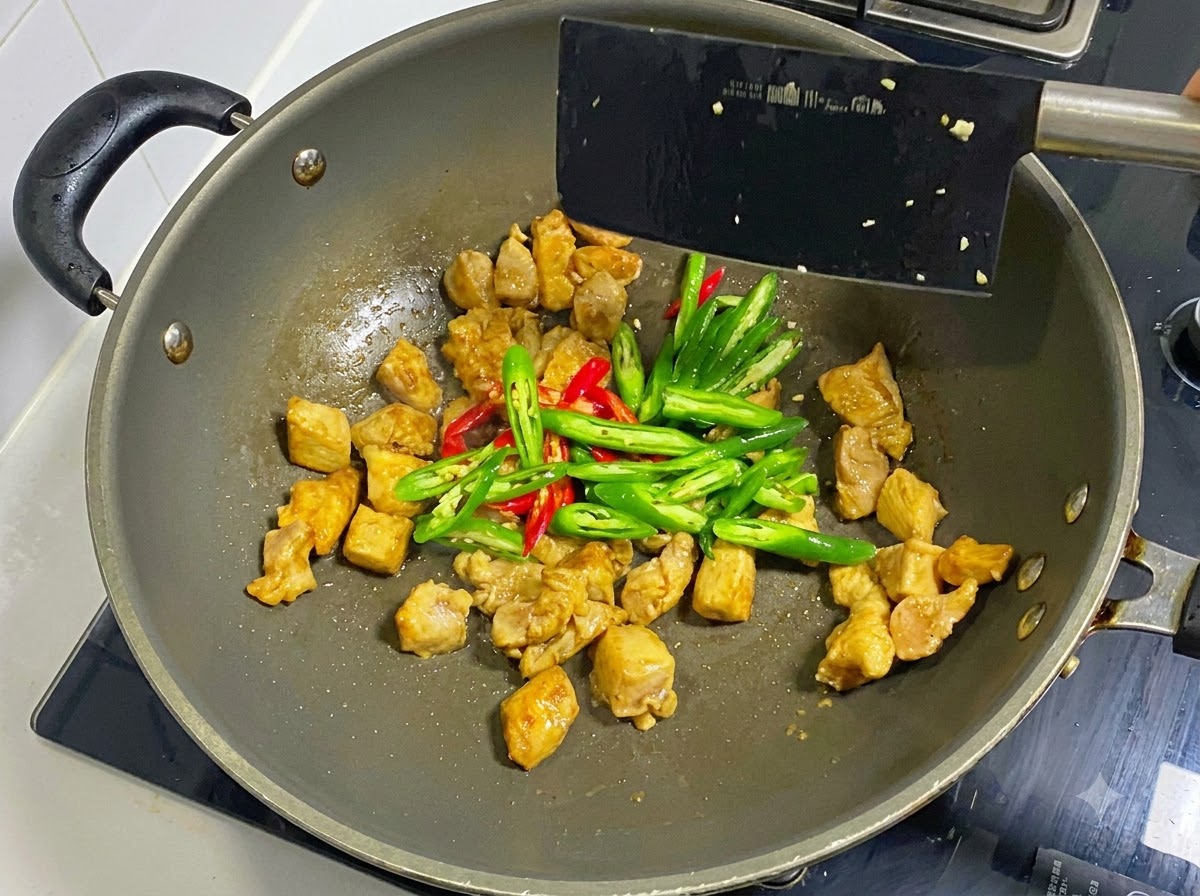 Pokrojone zielone i czerwone chili dodane do ugotowanych kawałków kurczaka w woku, mieszane czarną łopatką.