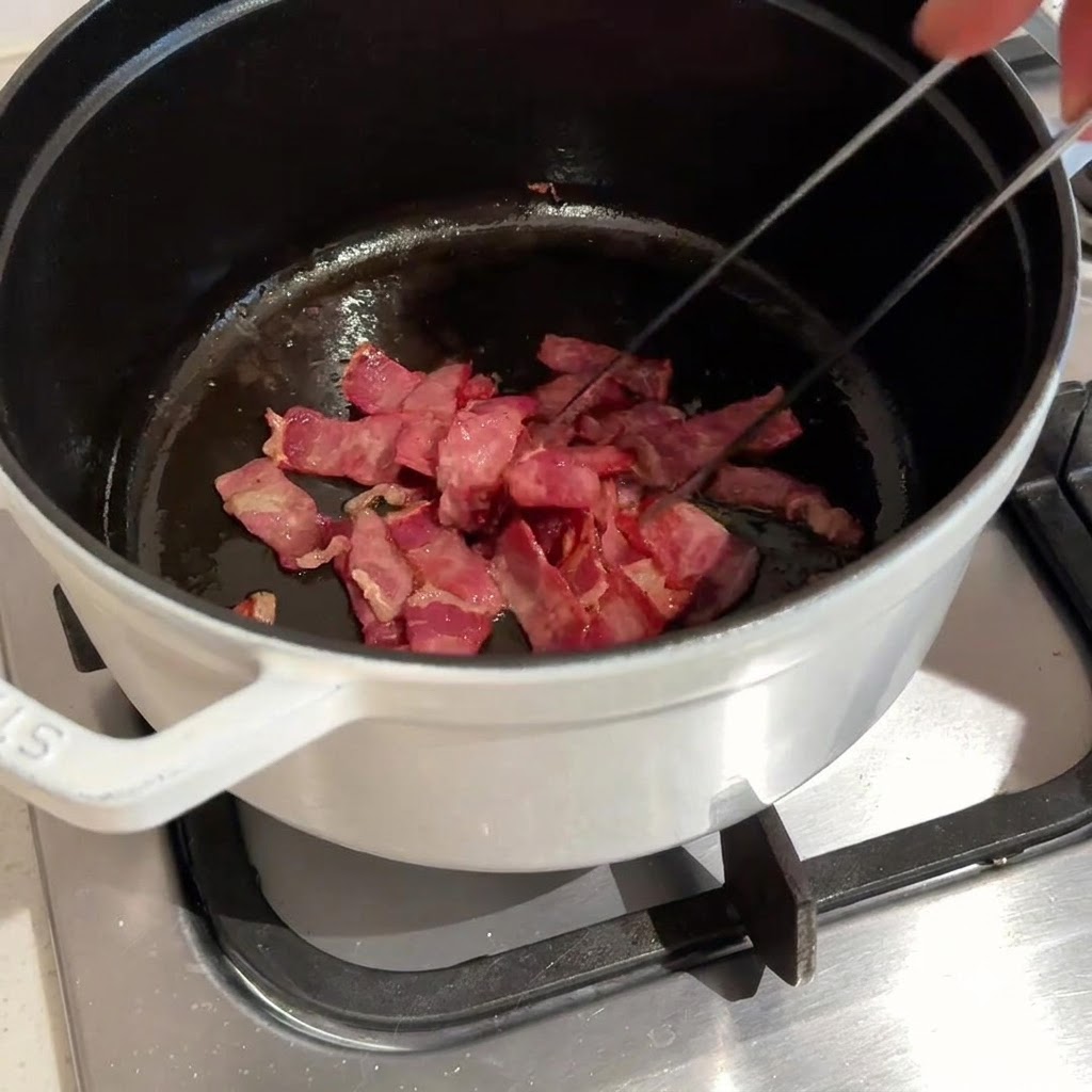 Posiekany boczek smażony i mieszany w ciemnym garnku za pomocą metalowych szczypiec.