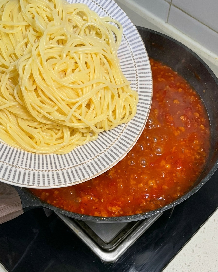 Świeżo ugotowane spaghetti przekładane z ozdobnego talerza na dużą patelnię z bulgoczącym, czerwonym sosem mięsnym.