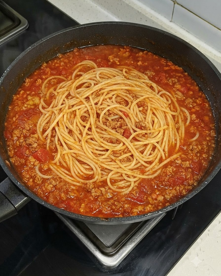 Gniazdo makaronu spaghetti równomiernie ułożone na dużej patelni wypełnionej czerwonym sosem mięsnym.