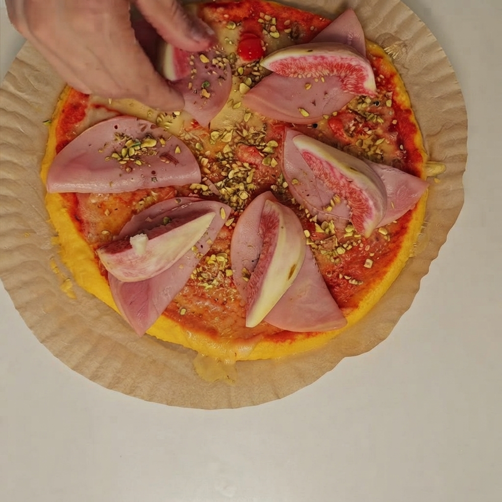 Osoba układająca świeże ćwiartki fig i pokruszone pistacje na plastrach szynki na pizzy.